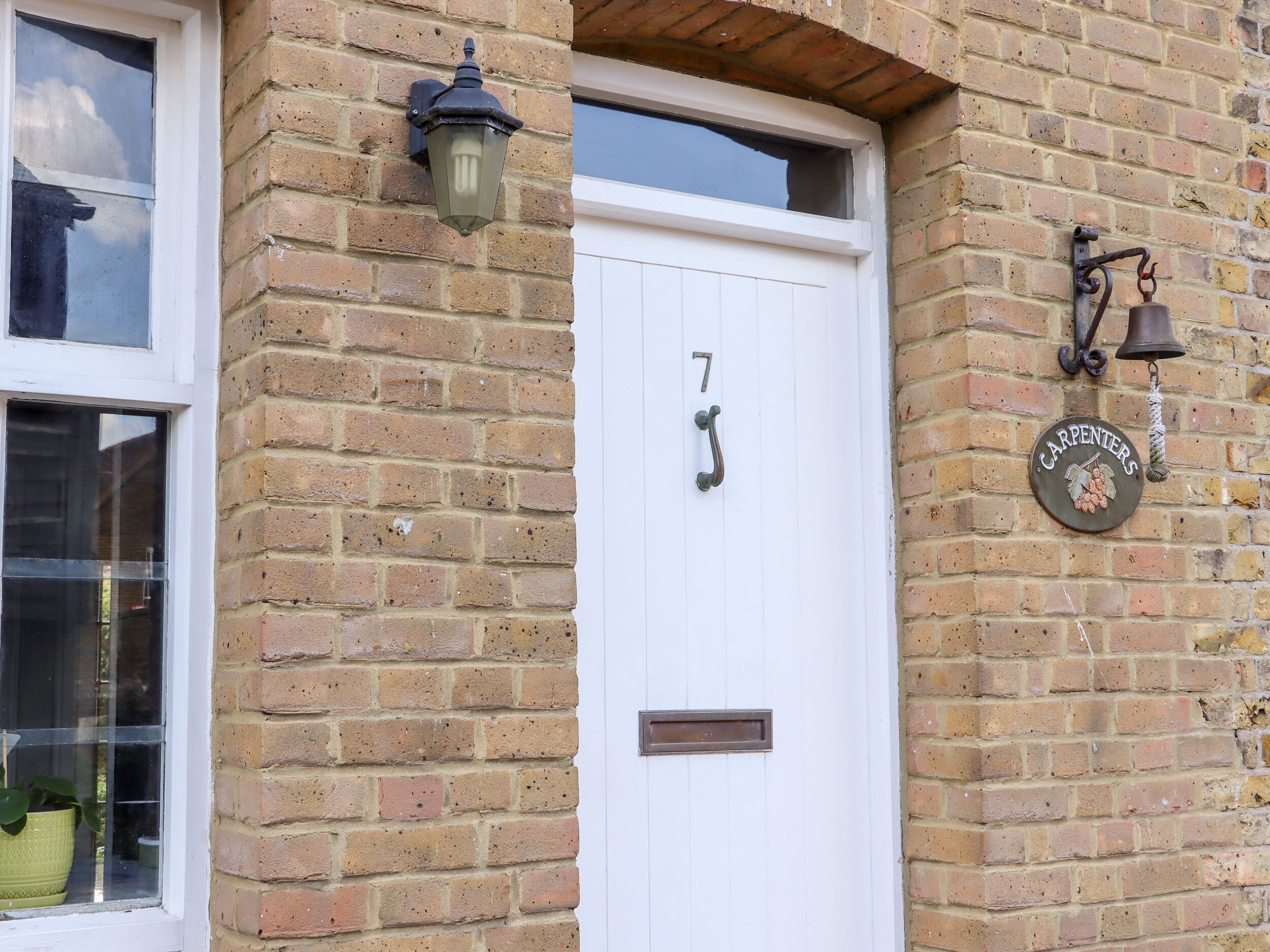 A brick building with a white door labeled "7", a window, a wall-mounted lantern, and a bell with a 'Carpenters' sign.