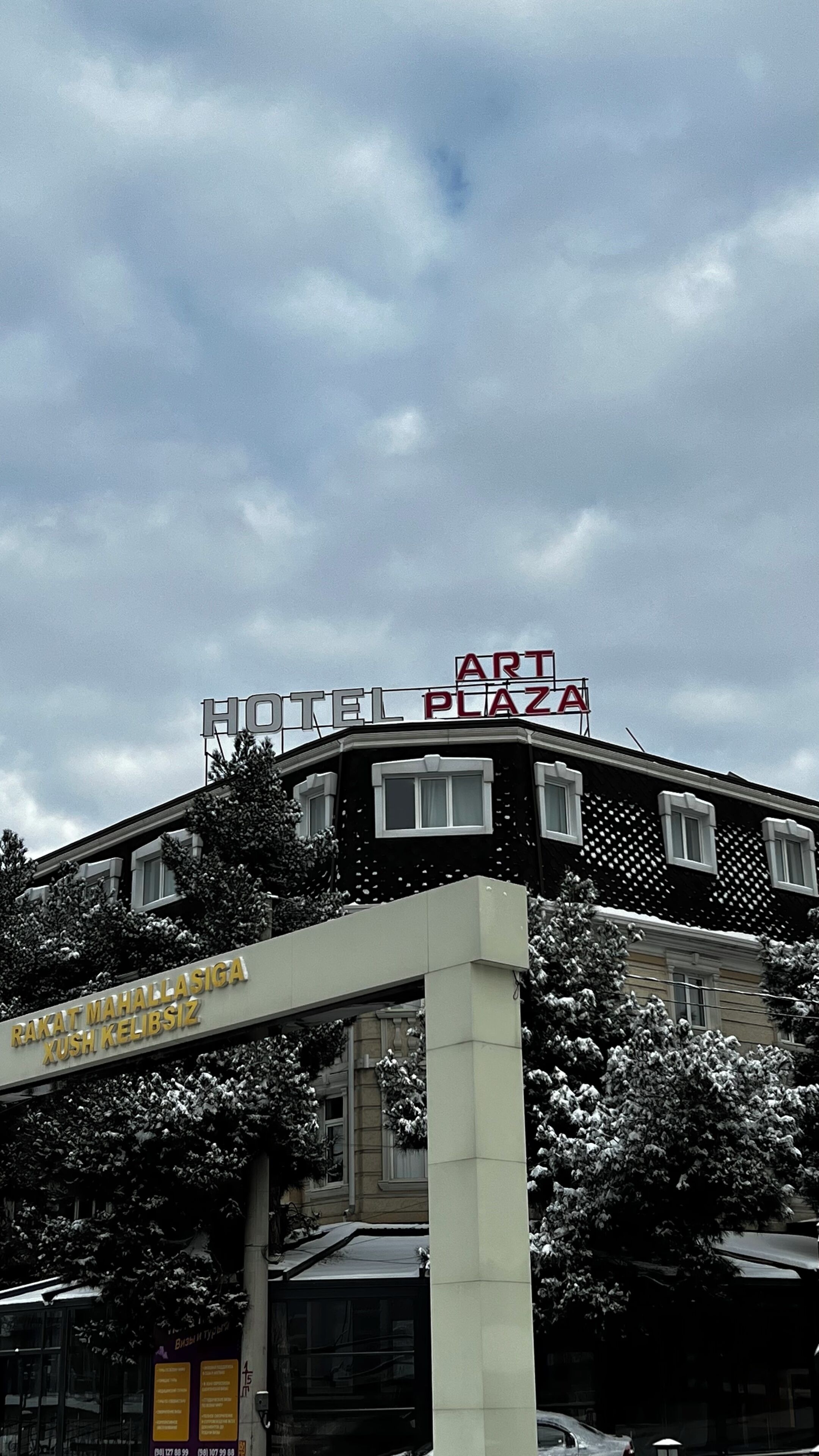 A hotel with a sign reading 'HOTEL PLAZA ART' and another sign below it that reads 'RAKAT MAHALLASIGA XUSH KELIBSIZ.'