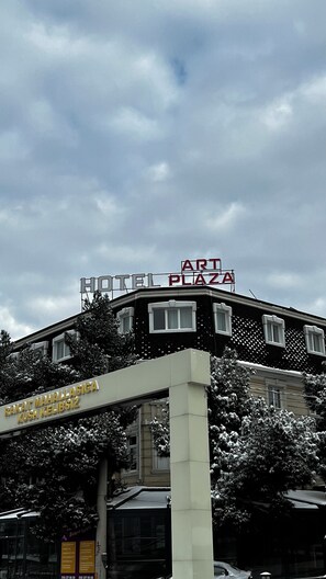 Um hotel com uma placa indicando 'HOTEL PLAZA ART' e outra placa abaixo com a inscrição 'RAKAT MAHALLASIGA XUSH KELIBSIZ'.