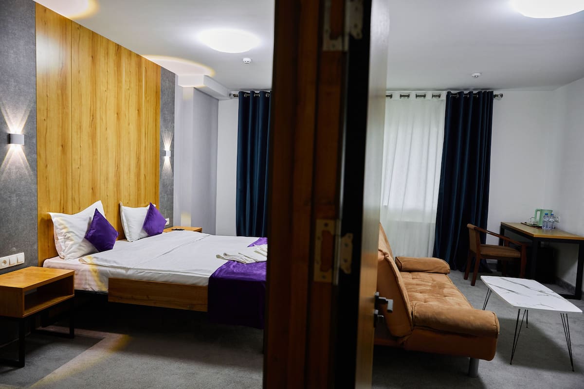 A hotel room with a wooden headboard, a bed with white and purple bedding, a brown sofa, a small table, and a desk with a chair.