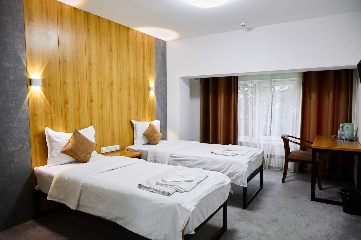 A hotel room with two beds, wooden headboard, white linens, a desk with a chair, and a window with curtains.