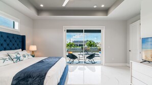 A bedroom with a bed, a nightstand, a lamp, a TV, and a sliding glass door leading to an outdoor patio with chairs. - Heated Pool and Spa Villa with Waterfront Views and Kayaks! (Cape Coral)