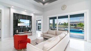 A modern living room with a large TV, a red chair, a beige sofa, and a view of a swimming pool. - Heated Pool and Spa Villa with Waterfront Views and Kayaks! (Cape Coral)