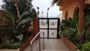 Une entrée fermée par un portail en métal, entourée d’une végétation luxuriante et bordée d’un chemin dallé menant à un bâtiment doté d’arcades.