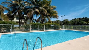 A rectangular swimming pool with clear blue water, surrounded by a white fence, palm trees, and a clear sky. - Cocon Dumyse, proche du Jardin botanique (Petit-Bourg)