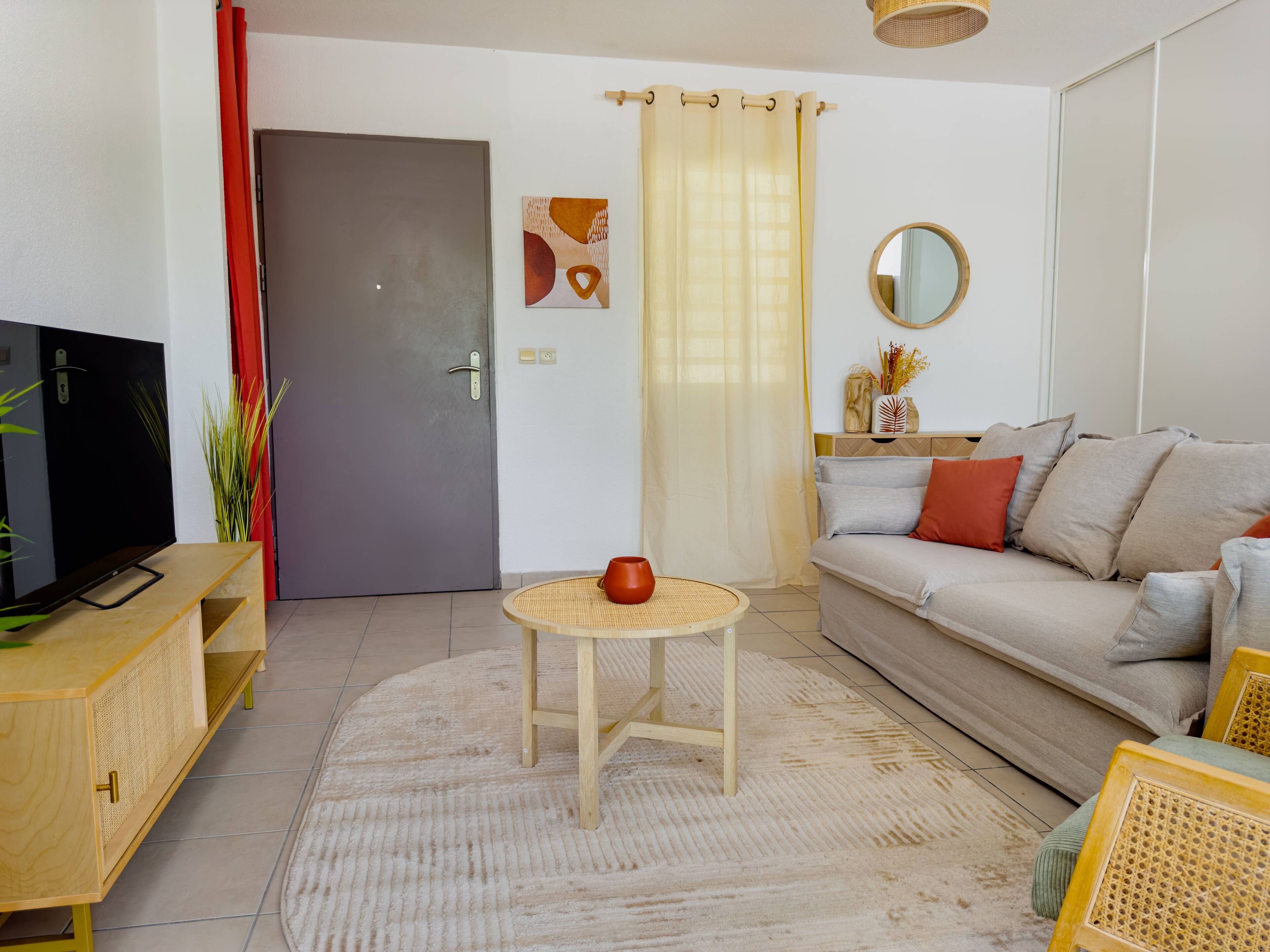 A living room with a sofa, a TV, a round table with a red pot, and a door with a red curtain.