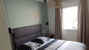 A bedroom with a bed, a bedside table, a hanging light, and a window with curtains. - Maison à 15 Minutes de la mer (Saint-Urbain)