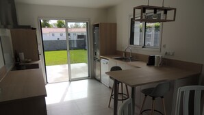 A modern kitchen with a large sliding glass door leading to a patio, a dining area with two bar stools, and a built-in oven. - Maison à 15 Minutes de la mer (Saint-Urbain)