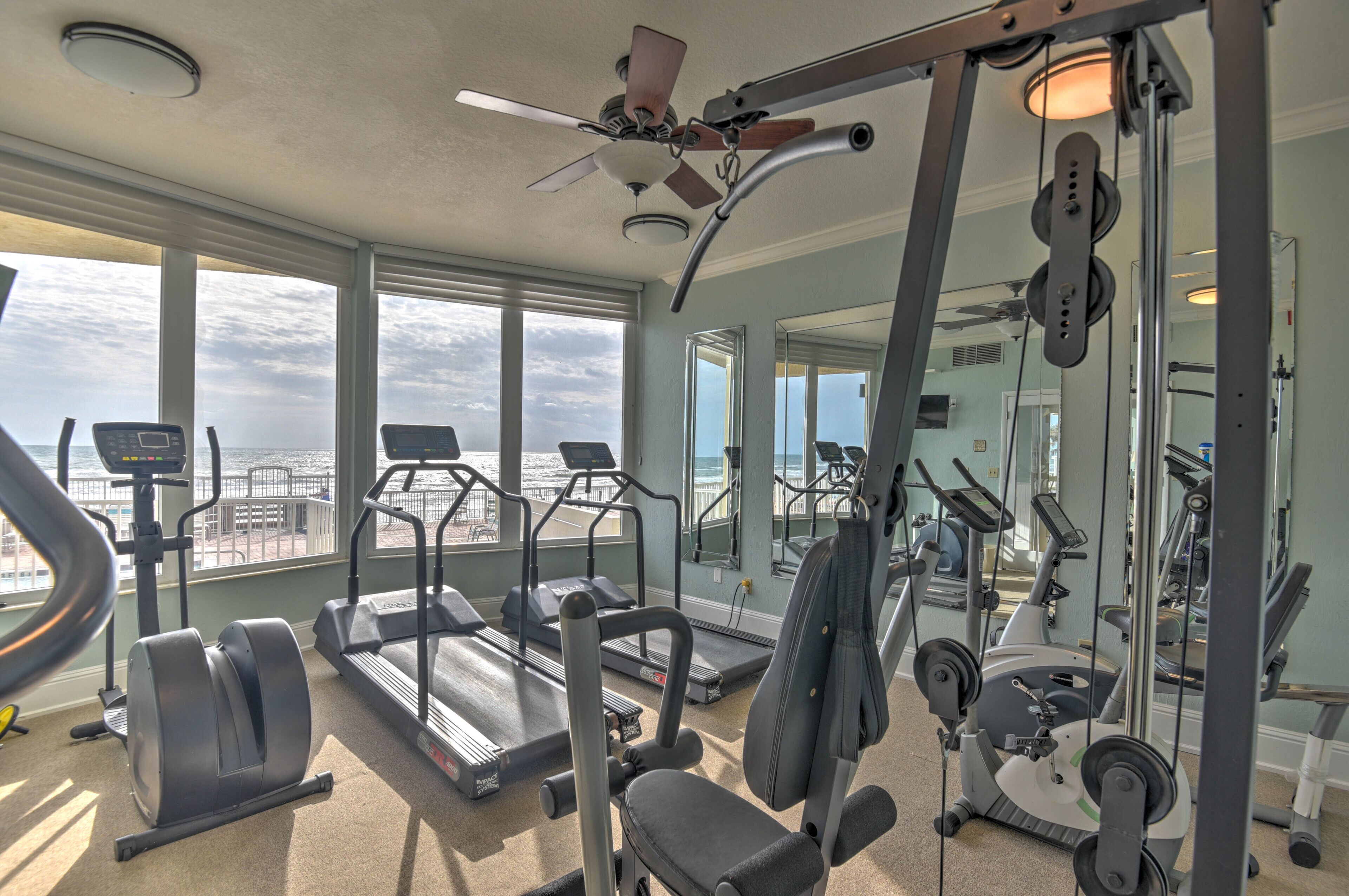 Un gimnasio con cintas de correr, elípticas y equipo de pesas, ventanas amplias con vista al océano y un ventilador de techo.