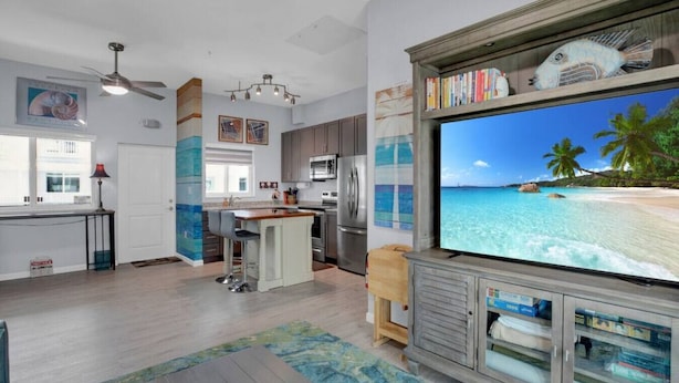 Una cocina moderna con una isla grande, una zona de comedor con taburetes de bar y una televisión grande mostrando una playa.