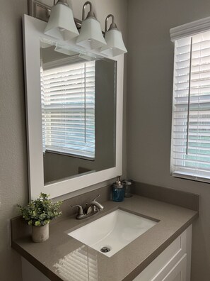 A bathroom with a white sink, a mirror with three lights, a potted plant, and a window with blinds. - Studio Apt. Near Downtown. Buffalo Bayou Park. (Houston)