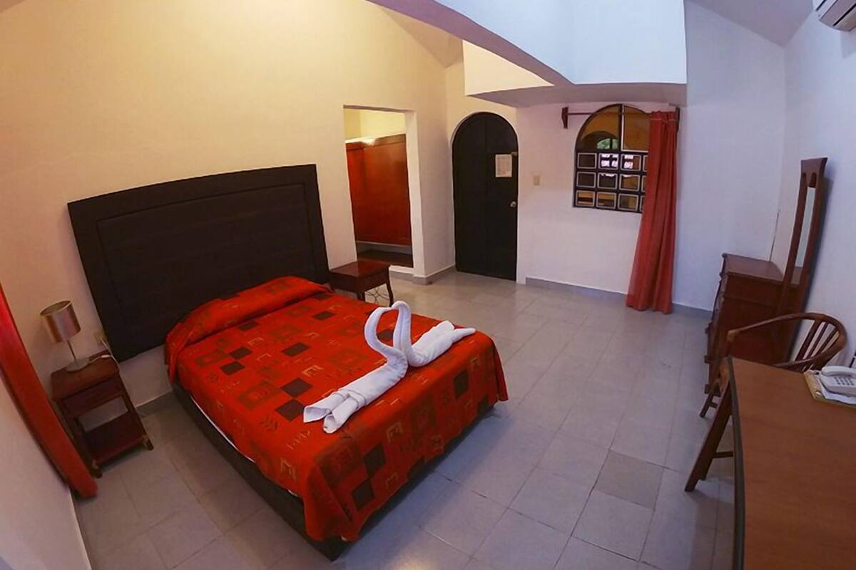 Quarto de hotel com uma cama grande, uma escrivaninha, uma cadeira, um guarda-roupa e uma porta que dá para outro cômodo.