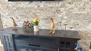A wooden entertainment center with a TV mounted above it, a fireplace with logs, and decorative items including wooden figures and a peacock.