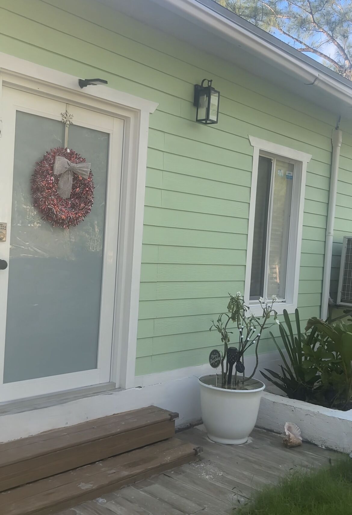 Een groen huis met een witte deur, een krans, een potplant en een kleine opstap naar de deur.