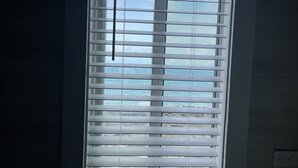 A window with horizontal blinds, a visible electrical outlet on the wall, and a view of the sky and clouds.