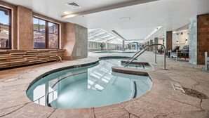 An indoor swimming pool with clear glass walls, stone flooring, and a seating area with benches.