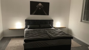 A modern bedroom with a large bed, two bedside lamps, a wall-mounted artwork, and a window with blinds.