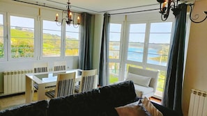 A room with a dining table and chairs, a sofa, and large windows with curtains. - Cortiguera Villas Lighthouse (Suances)