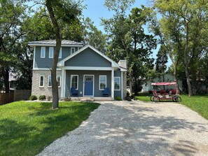 A two-story house with a front porch, a gravel driveway, and a red and white covered wagon in the background.