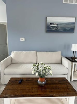 A living room with a white sofa, a wooden coffee table with a vase of flowers, and a painting on the wall.