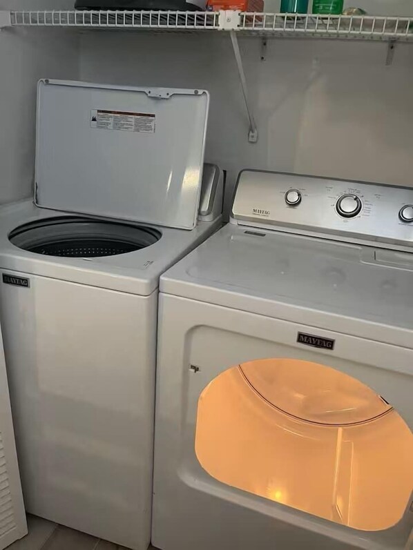 A compact laundry area with a front-loading washer and dryer, a shelf above, and a visible control panel.