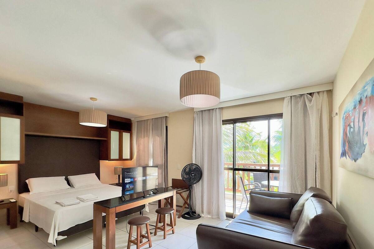 A hotel room with a bed, a desk with a computer, a fan, a couch, and a large window with curtains.