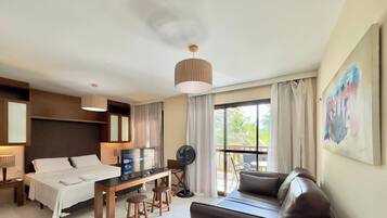 A hotel room with a bed, a desk with a computer, a fan, a couch, and a large window with curtains.