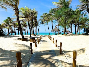 A wooden pathway leading to a beach with palm trees and thatched-roof structures. - Incrível Resort VG Sun (Caucaia)