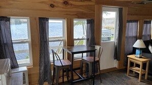 Una habitación con una mesa y sillas de madera, una mesita con lámpara y ventanas con cortinas que dan a un cuerpo de agua.