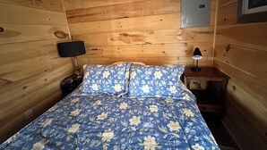 Una habitación de cabaña de madera con una cama, dos lámparas de noche, una mesita de noche y un cuadro eléctrico en la pared.