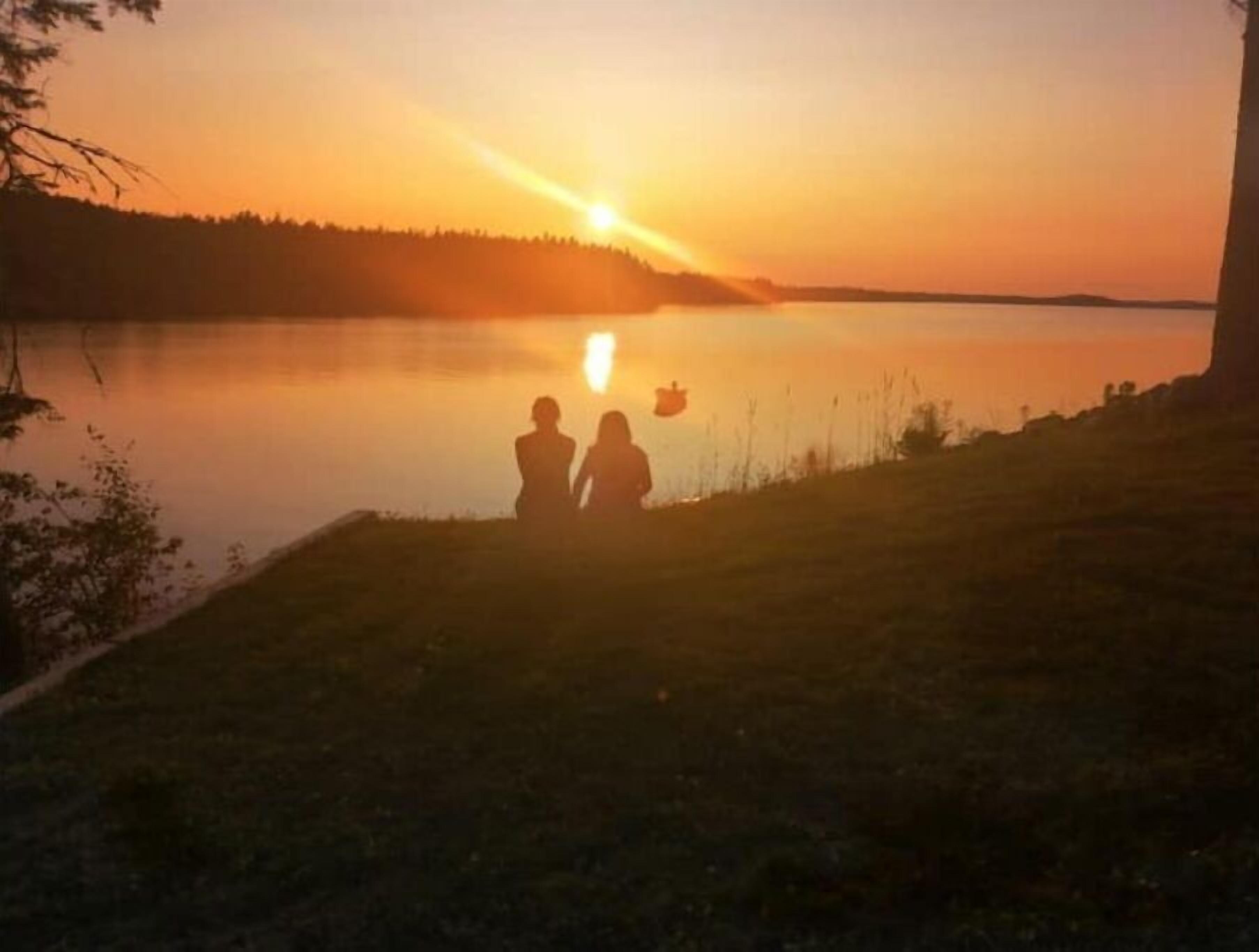 A sunset over a calm lake with two silhouettes of people standing on the shore.