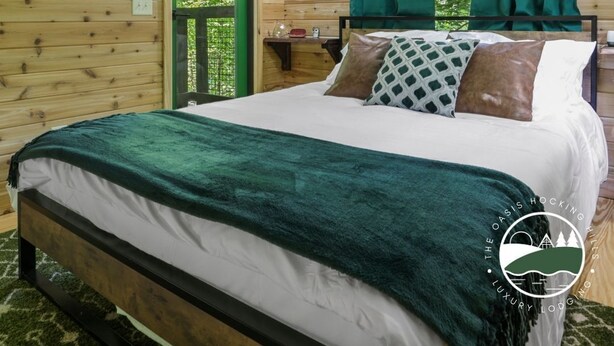 A wooden cabin bedroom with a bed, green curtains, and a window with a view of greenery. - Rustic Treehouse Oasis in Hocking  Hills --Adventure Awaits (Logan)