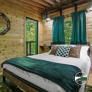 A wooden cabin bedroom with a bed, green curtains, and a window with a view of greenery. - Rustic Treehouse Oasis in Hocking Hills --Adventure Awaits (Logan)