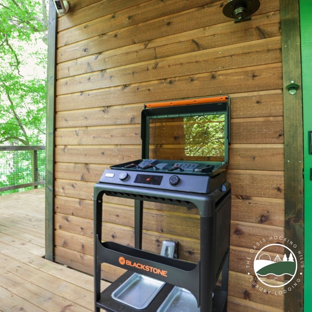 A portable gas stove with a built-in grill, placed on a wooden deck against a wooden wall.