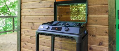 A portable gas stove with a built-in grill, placed on a wooden deck against a wooden wall.