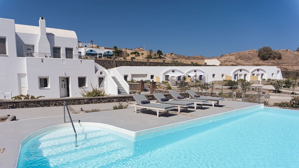 A pool area with lounge chairs and a clear blue pool, surrounded by white buildings with arched windows and a stone wall. - VILLAS SCIROCCO (Santorini)