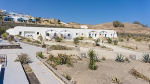A white building with multiple arched doorways, surrounded by desert vegetation and a hilly landscape. - VILLAS SCIROCCO (Santorini)