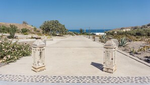 A paved pathway leading to a gate with two stone pillars, surrounded by desert vegetation and a clear view of the sea. - VILLAS SCIROCCO (Santorini)