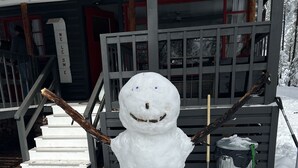 Un muñeco de nieve con ramas por brazos, situado frente a una casa con porche y escalones.