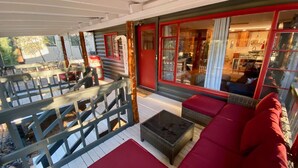 Un rincón exterior acogedor con un sofá rojo y una mesita, una terraza de madera, y vistas del interior a través de amplios ventanales.