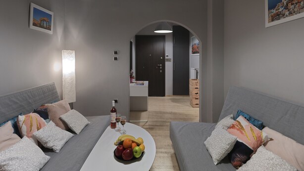 Uma sala de estar moderna com sofás cinzentos, uma mesa de centro branca com frutas e copos, e uma porta escura que dá para uma cozinha.