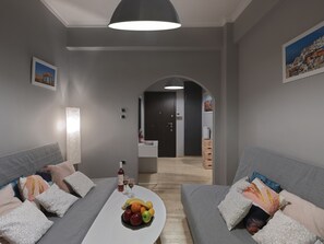 A modern living room with grey sofas, a white coffee table with fruits and glasses, and a dark door leading to a kitchen. - Makrigianni19• Comfortable apartment opposite Acro (Athens)