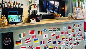 Zona de bar con pantalla digital, una variedad de botellas, un tablero de menú y una pared decorada con banderas.