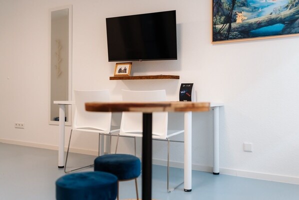 A modern room with a flat-screen TV mounted on the wall, a small wooden table, white chairs, and a blue ottoman. - Hostel Mannheim (Mannheim)