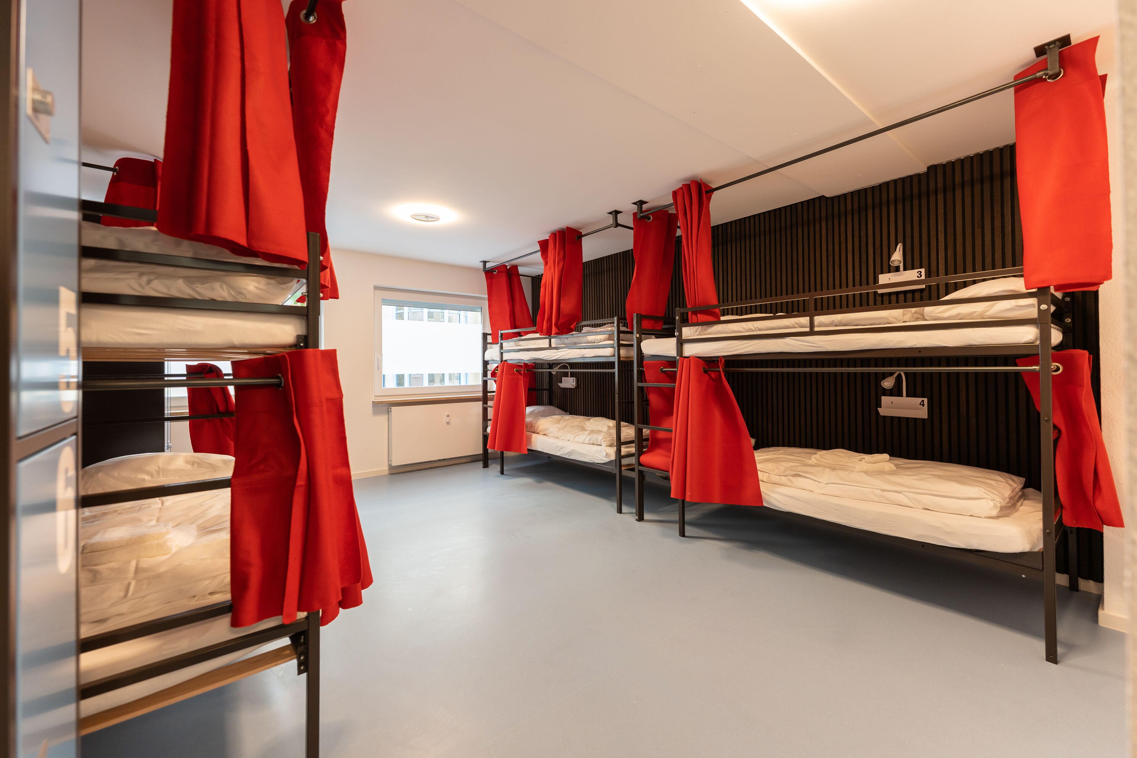 A dormitory room with bunk beds, red curtains, and a window with blinds.