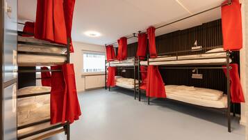 A dormitory room with bunk beds, red curtains, and a window with blinds.