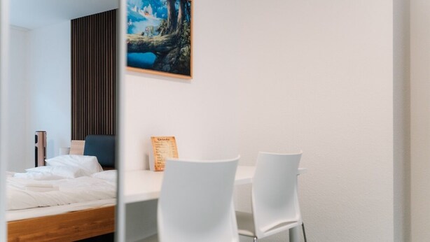 A modern hotel room with a wooden bed, white chairs, a dining table, and a painting on the wall.