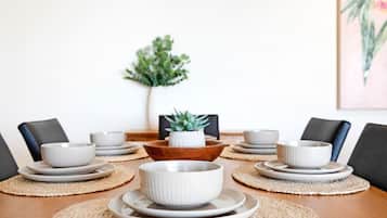 A dining table set with white bowls and plates, surrounded by chairs, with a plant and a framed picture on the wall.