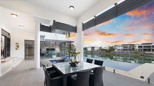 A modern dining area with a glass table and black wicker chairs, overlooking a body of water and a sunset view.