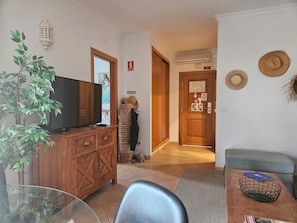 A living room with a TV, wooden cabinet, and a plant. There's a hallway leading to a door with a sign.